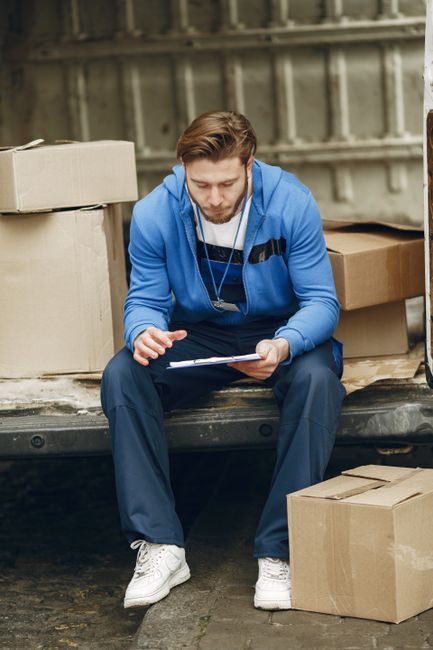 A man with light skin and brown hair, wearing a blue hoodie and dark trousers, is sitting on the edge of an open van in an outdoor setting during daytime. He is holding a clipboard and pen, appearing 
