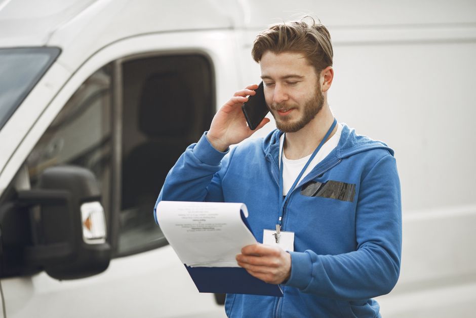 A man with light skin and brown hair, wearing a blue hoodie and dark trousers, is sitting on the edge of an open van in an outdoor setting during daytime. He is holding a clipboard and pen, appearing 