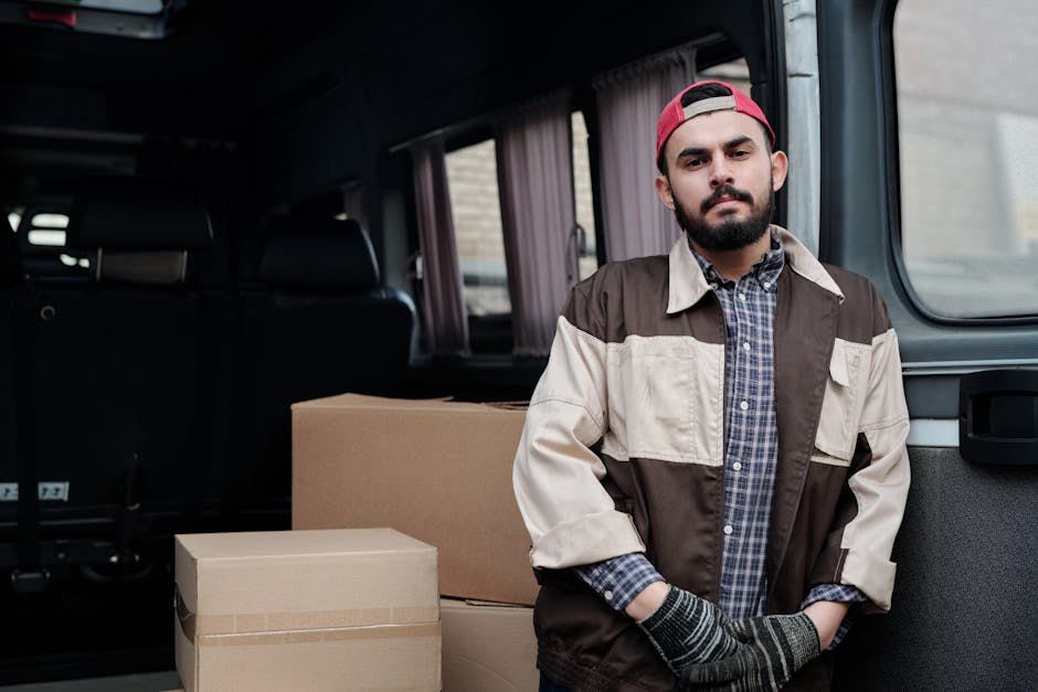 A man standing beside an open vehicle, likely a van used for home relocation, inside a residential area with a brick building visible in the background. He is wearing a beige, brown, and white work jacket over a checked shirt, with black gloves and a red cap worn backwards. The vehicle's interior, visible behind him, contains large cardboard boxes ready for packing and moving furniture. The boxes are stacked on the vehicle's floor, some sealed with packing tape, indicating preparation for loading or unloading. The ambiance suggests a professional removal service, with the man representing [COMPANY_NAME] during the furniture transport and packing process associated with house removals near Ealing Broadway station. The scene emphasizes the logistics of loading a van used in local moves, with soft natural lighting highlighting the packing materials and the worker's focused expression.