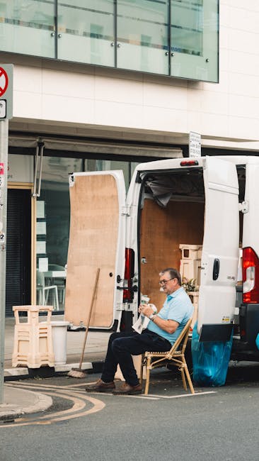A man with glasses wearing a light blue shirt sits on a wooden chair parked outside a white delivery van during a house removal process. The van's rear doors are open, revealing a partially loaded interior with large cardboard boxes and wrapped furniture for a home relocation. A wooden panel is propped against the van on the pavement, indicating ongoing packing or loading activities. Nearby, a wooden shelving unit or small table is visible, and the scene is set against a modern building with glass windows and a white exterior. The environment appears to be a quiet urban street with a pedestrian crossing and road markings visible. The man appears to be taking a break or reviewing documents related to the moving logistics, and the setting suggests a professional moving service, such as Man with Van Ealing, preparing for furniture transport and packing to facilitate a smooth house or flat removal near Ealing Broadway station.