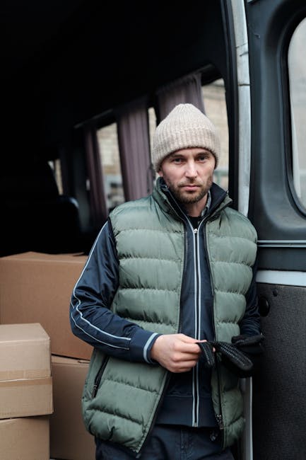 A man standing beside an open vehicle, likely a van used for home relocation, inside a residential area with a brick building visible in the background. He is wearing a beige, brown, and white work jacket over a checked shirt, with black gloves and a red cap worn backwards. The vehicle's interior, visible behind him, contains large cardboard boxes ready for packing and moving furniture. The boxes are stacked on the vehicle's floor, some sealed with packing tape, indicating preparation for loading or unloading. The ambiance suggests a professional removal service, with the man representing [COMPANY_NAME] during the furniture transport and packing process associated with house removals near Ealing Broadway station. The scene emphasizes the logistics of loading a van used in local moves, with soft natural lighting highlighting the packing materials and the worker's focused expression.