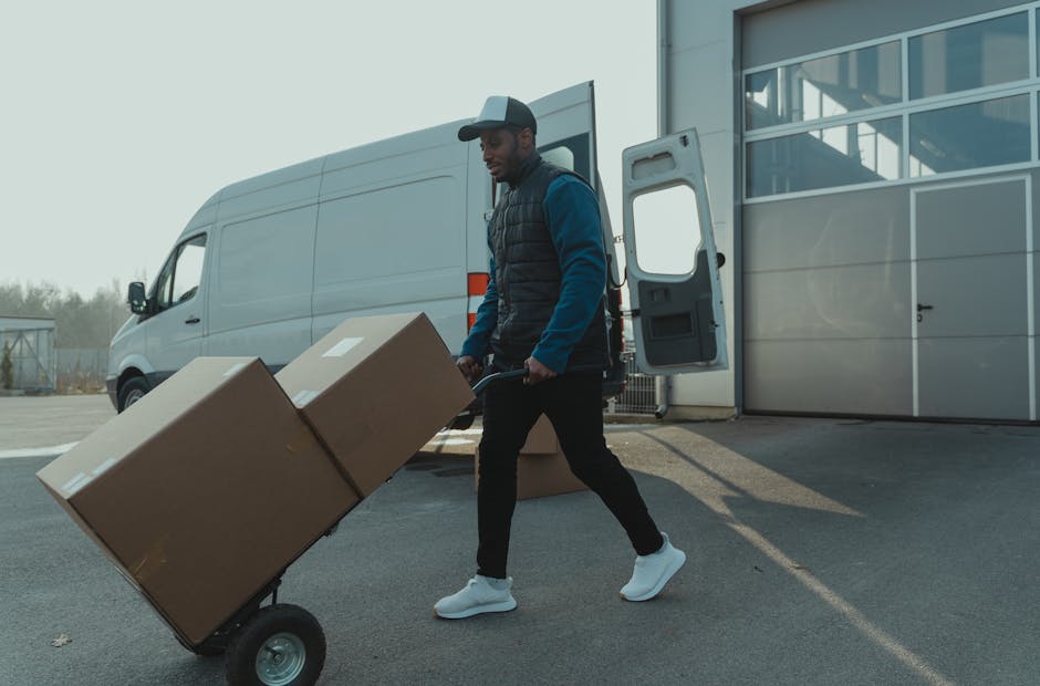 A man wearing a grey cap, blue long-sleeved shirt, and black puffer vest is seen pushing a hand truck loaded with two large cardboard boxes outside a white van, which is parked near a modern building with glass windows. The scene takes place on a paved area adjacent to the building, with the van's rear doors open, revealing an empty interior suitable for home relocation or furniture transport. The man appears to be engaged in a loading process, preparing for a house removal or moving service, with additional cardboard boxes lying on the ground nearby. The background includes a partly cloudy sky, and the overall environment suggests an organized move, facilitated by a professional removals service such as Man with Van Ealing, specialising in narrow street and terrace moves.