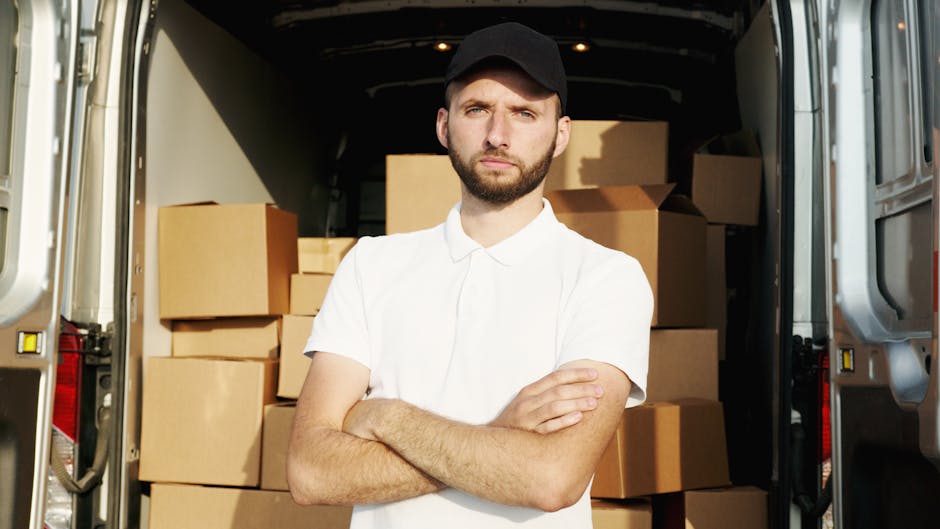 Two men are engaged in a home relocation task outside a residential property, preparing to load cardboard boxes into the back of a white van equipped for furniture transport. The man on the left, wearing a black vest over a blue long sleeve shirt, black gloves, and white sneakers, is holding a large cardboard box. The man on the right, dressed in a navy jacket, dark jeans, and white sneakers, is assisting with guiding the box onto the van's open rear. Nearby, there is a black hand truck with two wheels standing on a brick-paved driveway, ready to support the movement of multiple boxes. The open van interior is partially visible, showing numerous packed cardboard boxes of various sizes, some sealed with packing tape. The setting appears to be a cloudy day, with a brick wall, hedge, and residential rooftops in the background, illustrating a typical house move process that Man with Van Ealing may facilitate for typical home relocations involving narrow streets and terrace properties.