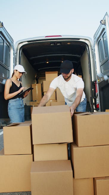 A man wearing a blue jacket, black padded vest, and a light grey cap is standing outdoors during daylight, handling cardboard boxes used for home relocation. He is gripping a hand truck or trolley, po