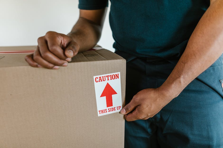 A person wearing a dark green T-shirt and blue work trousers is holding a medium-sized cardboard box with both hands. The box has a red and white warning label attached to the front that reads 'Caution' with an upward-pointing arrow and the instruction 'This side up.' The individual appears to be engaged in a home relocation process, possibly preparing the box for transport. Inside a well-lit indoor space, they are either about to place the box into a vehicle or are in the process of stacking or organizing packing materials. The cardboard box is sealed with packing tape, indicating careful packing and secure packaging typical of furniture transport and packing during a moving or removal service. This image exemplifies the meticulous handling involved in house removals, highlighting the importance of proper packing labels to ensure safe furniture transport and efficient loading in a professional moving context, such as those offered by Man with Van Ealing.