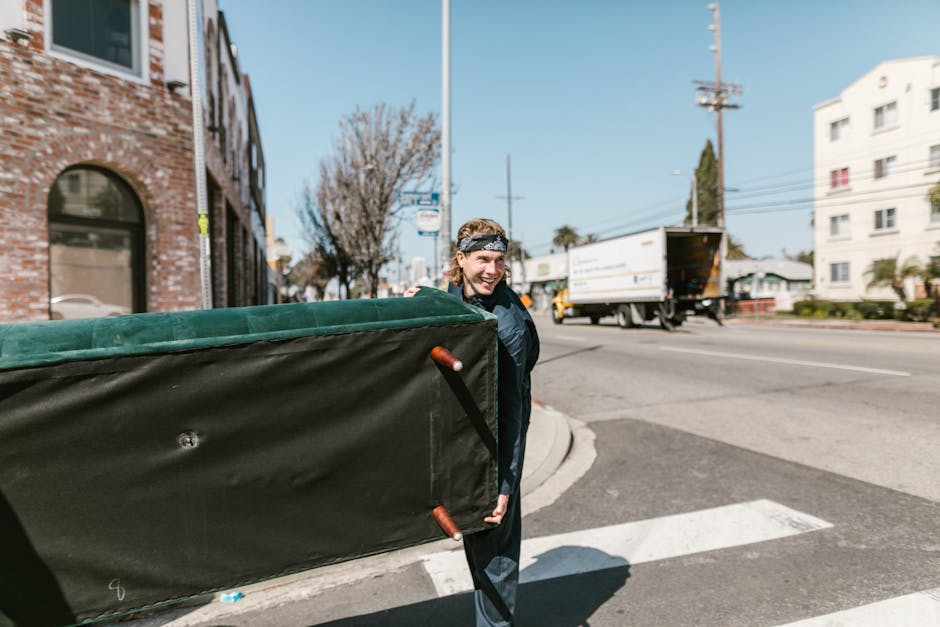A man wearing a black jacket and a patterned headband is outside on a city street, carrying a large, rectangular piece of furniture wrapped in green fabric and black protective material with brown leather handles. He is smiling as he lifts the item, which appears to be part of a home relocation process. Behind him, a white commercial van is parked near a building with arched windows, while a smaller delivery truck is seen driving in the background. The sidewalk is paved and the street has markings, with utility poles and trees lining the road. The scene is well-lit with natural daylight, indicating a clear day. This image illustrates the logistics involved in packing and loading furniture during house removals, relevant to services provided by Man with Van Ealing for W5 postcode relocations, as part of professional furniture transport and moving logistics.